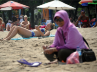 A Muslim woman wearing a veil sits in front of foreign tourists wearing bikinis on Kuta beach in Bali on June 6, 2013. Contestants at this year's Miss World beauty pageant will not wear bikinis in the parade in a bid to avoid causing offence in Muslim-majority Indonesia, organisers said on June 5, 2013. AFP PHOTO/SONNY TUMBELAKA (Photo credit should read SONNY TUMBELAKA/AFP/Getty Images)