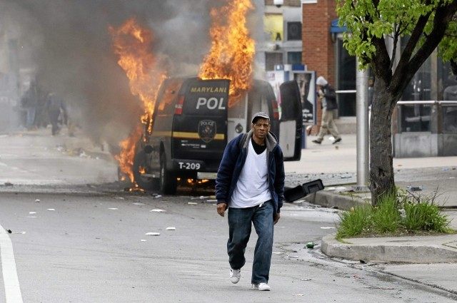 A man walks past a burning police vehicle, Monday, April 27, 2015, during unrest following