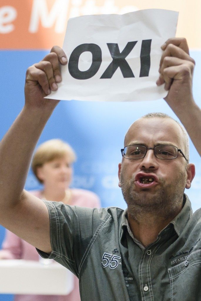 A protester holds a sheet with the Greek word for 'No' during an open house presentation of Germany's conservative Christian Democratic Union (CDU) while German Chancellor Angela Merkel speaks in the background. AFP PHOTO / CLEMENS BILAN