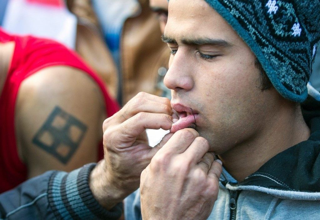 A man uses needle and thread to sew shut the mouth of a compatriot from Iran as a sign of protest as migrants and refugees wait to cross the Greek-Macedonian border near Gevgelija on November 26, 2015. Since last week, Macedonia has restricted passage to northern Europe to only Syrians, Iraqis and Afghans who are considered war refugees. All other nationalities are deemed economic migrants and told to turn back. Over 1,500 people are stuck on the border, mostly Indian, Moroccan, Bangladeshi and Pakistani. AFP PHOTO / ROBERT ATANASOVSKI / AFP / ROBERT ATANASOVSKI (Photo credit should read ROBERT ATANASOVSKI/AFP/Getty Images)