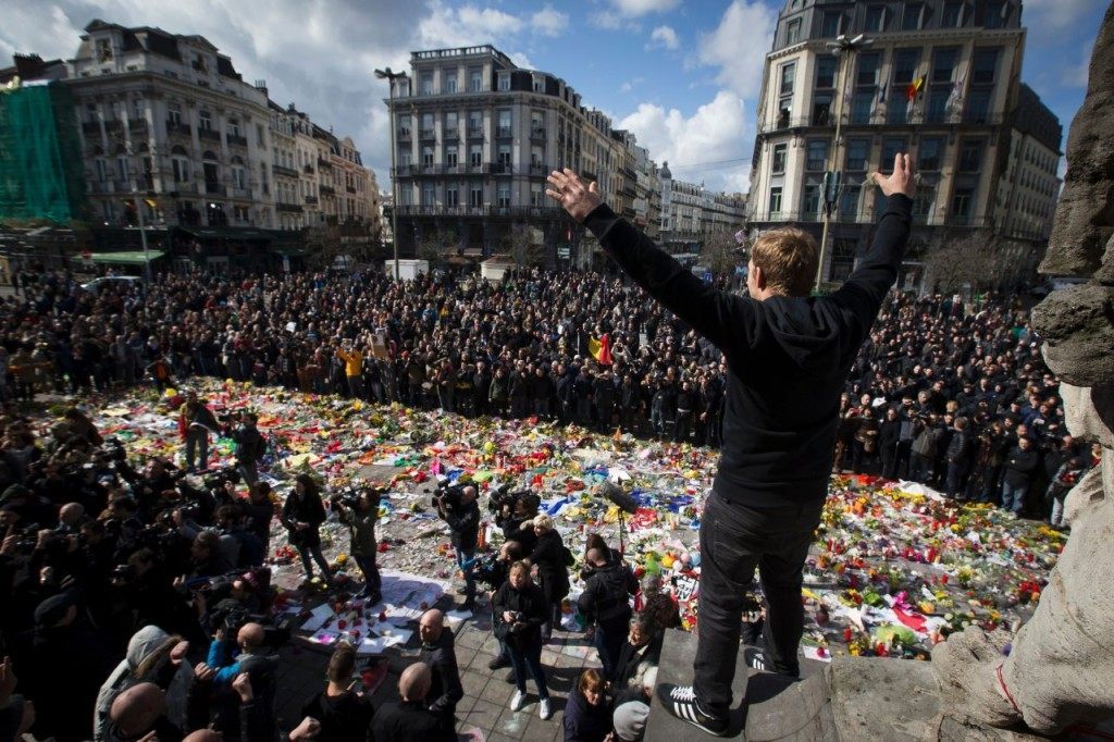 TOPSHOT - A man gestures as masked and hooded men arrive outside the stock exchange in Brussels on March 27, 2016 as tensions mounted after the square was invaded by some 200 far-right football hooligans. Police fired water a cannon at far-right football hooligans who invaded a square in the Belgian capital that has become a memorial to the victims of the Brussels attacks, an AFP journalist said. Police took action after about 200 black-clad hooligans shouting nationalist and anti-immigrant slogans moved in on the Place de la Bourse where people were gathering in a show of solidarity with the victims. / AFP / Belga / KRISTOF VAN ACCOM / Belgium OUT (Photo credit should read KRISTOF VAN ACCOM/AFP/Getty Images)