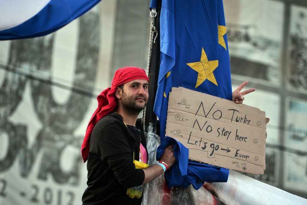 A man holds a placard as he stands next to an EU flag at the port at the town of Chios where refugees and migrants who broke out from Vial detention center are camped out, on April 3, 2016. Under a European Union deal with Turkey, migrants and refugees arriving after March 20 are to be held in centres set up on five Aegean islands, including Chios, and sent back if their asylum applications are not accepted. Returns are supposed to begin on April 4 / AFP / LOUISA GOULIAMAKI (Photo credit should read LOUISA GOULIAMAKI/AFP/Getty Images)