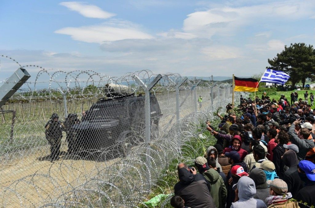 TOPSHOT - Refugees and migrants face Macedonian soldiers at the border fence with Greece and German flags as they protest to reopen the border near their makeshift camp in the northern border village of Idomeni, Greece, on April 11, 2016. Fresh protests broke out as Greece and Macedonia blamed each other for an incident in which tear gas and rubber bullets were fired on migrants trying to breach the closed border to get to western Europe on April 10. Medical charity Doctors Without Borders (MSF) said 260 people were treated for injuries after the flare-up on the Greece-Macedonia border. / AFP / BULENT KILIC (Photo credit should read BULENT KILIC/AFP/Getty Images)