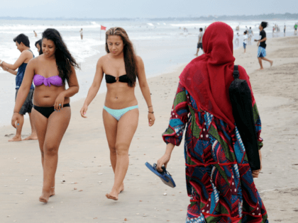 A Muslim woman (R) wearing a veil walks past foreign tourists wearing bikinis on Kuta beach near Denpasar on Indonesia's resort island of Bali on June 6, 2013. Contestants at this year's Miss World beauty pageant will not wear bikinis in the parade in a bid to avoid causing offence …