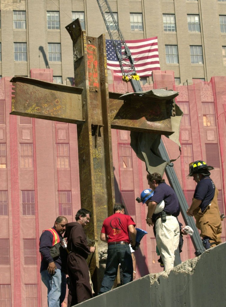 Father Brian Jordan, second from left, blesses, Thursday, Oct. 4, 2001, a cross of steel beams found amidst the rubble of the World Trade Center by a laborer two days after the collapse of the twin towers. The cross was from World Trade tower One, and was found in World Trade building Six and moved to its present location Wednesday. Other rescue and construction workers join Jordan for the ceremony. A protective mesh hangs on the building in the background. (AP Photo/Pool, Kathy Willens)