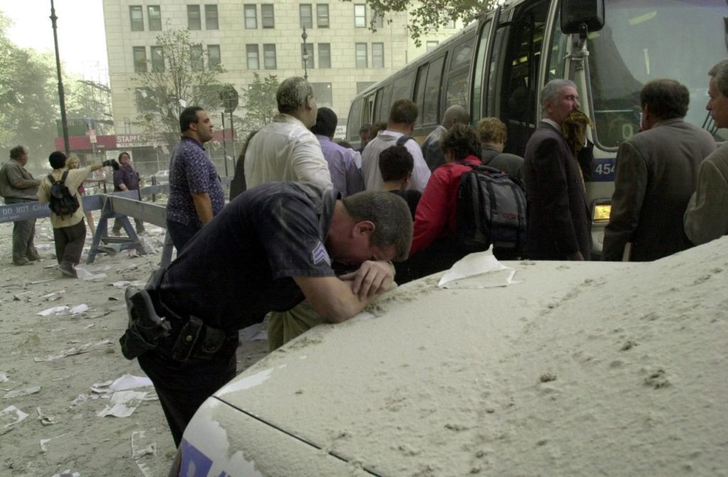 An exausted police officer rests on a car covered in dust near the World Trade Center 11 September 2001 in New York as people board a bus to be evacuated after two hijacked planes crashed into the landmark towers. AFP PHOTO/Stan HONDA / AFP / STAN HONDA (Photo credit should read STAN HONDA/AFP/Getty Images)