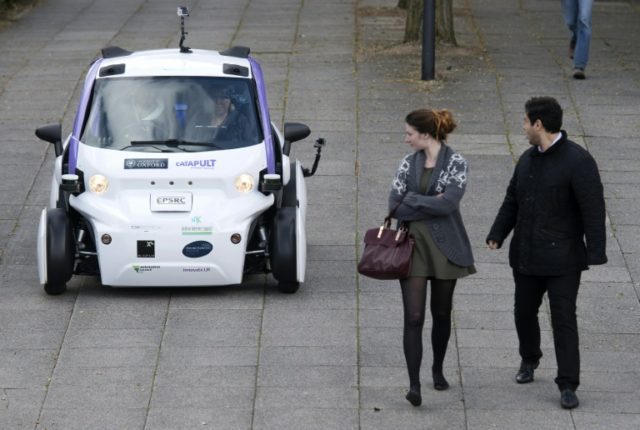 Bystanders look at an autonomous self-driving vehicle as it is tested in a pedestrianised