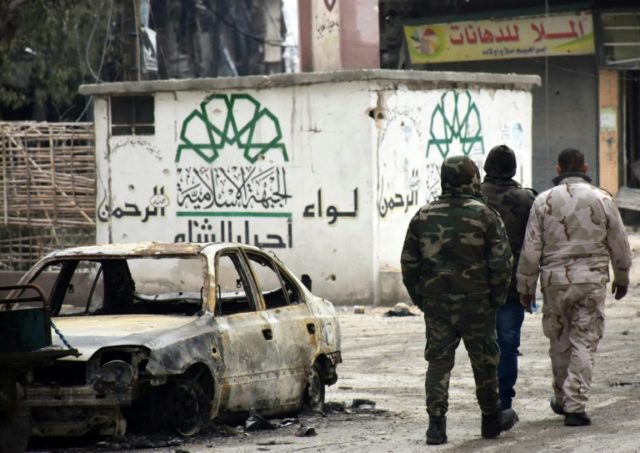 Syrian government forces walk past a destroyed vehicle and a graffiti bearing the emblem o