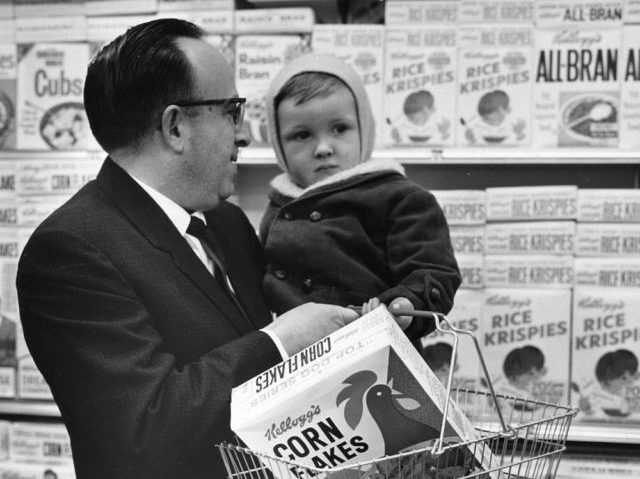 4th March 1974: A father holds his baby on one arm and the Kellogg's Cornflakes on the other, whilst shopping at a Foodtown Supermarket in Lewisham, London. (Photo by Evening Standard/Getty Images)