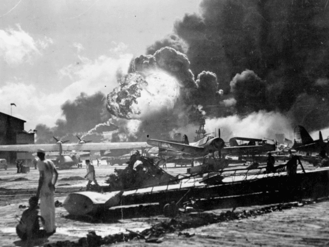 An explosion at the Naval Air Station, Ford Island, Pearl Harbour (Pearl Harbor) during the Japanese attack. Sailors stand amid wrecked watching as the USS Shaw explodes in the center background. The USS Nevada is also visible in the middle background, with her bow headed toward the left. (Photo by …