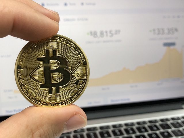 Bitcoin held in front of a computer screen showing a price chart on Coinbase.