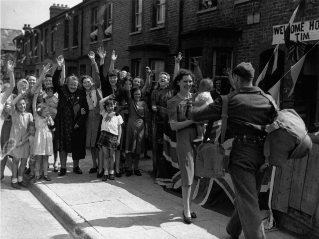 1944: Neighbours wave and cheer as they watch a young soldier being welcomed home from the Second World War by his wife and baby.