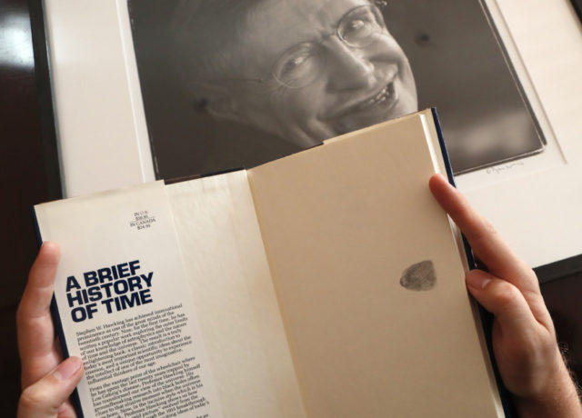 Britain Stephen Hawking Auction A Book, signed with a thumb print by Stephen Hawking is one of the personal and academic possessions of Stephen Hawking, photo behind, at the auction house Christies in London, Friday, Oct. 19, 2018. The online auction announced Monday Oct. 22, 2018, by auctioneer Christie’s features 22 items from Hawking, including his doctoral thesis on the origins of the universe, with the sale scheduled for 31 October and 8 November. (AP Photo/Frank Augstein) The Associated Press