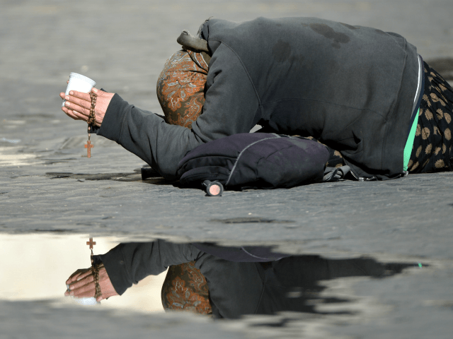 A beggar lies on the ground asking for some money, while holding a rosary, in a street of