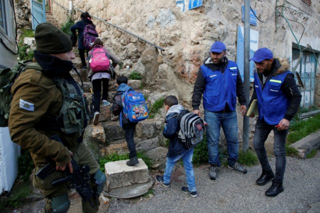 An Israeli soldier (L) stands guard as members of the Palestinian Youth Against Settlements (YAS) activists escort children on their way to school in the occupied West Bank town of Hebron on February 10, 2019 Palestinians walk Hebron pupils to school after monitors quit