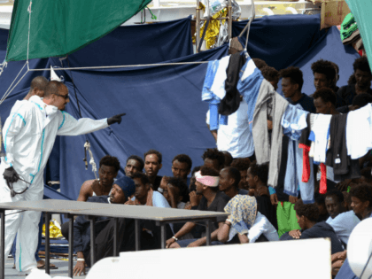 An official wearing a protective suit gestures towards migrants as they sit on the deck of the Italian Coast Guard vessel "Diciotti" in the Sicilian port of Catania, on August 23, 2018, as they wait to disembark following a rescue operation at sea. - The Diciotti vessel rescued the migrants …