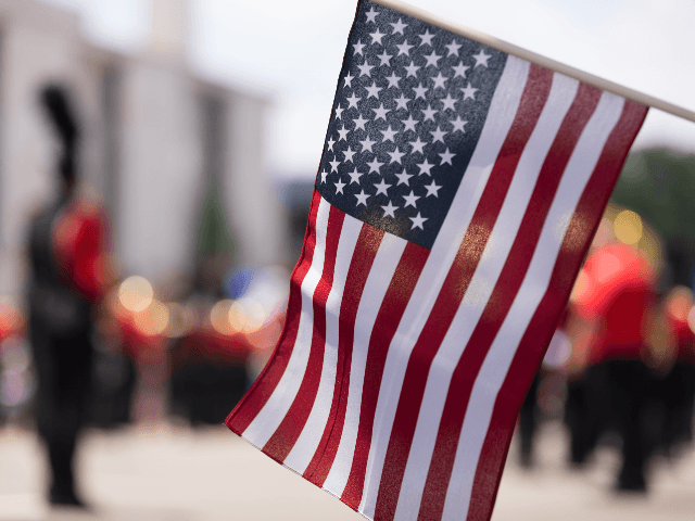 The American National Flag on the foreground with a marching band and buildings out of focus during a parade.