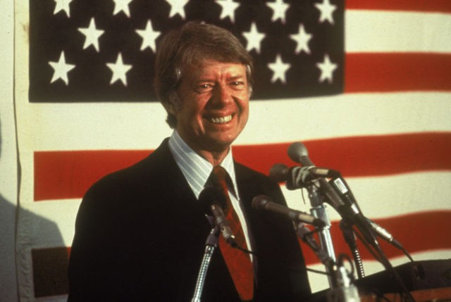 U.S. president Jimmy Carter smiling at a podium in front of an American flag, 1970s. (Phot