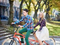 Young active happy traveler couple, handsome bearded man and pretty blond woman in glasses on tandem bike on bright sunny day on stony staircase and ancient building background.
