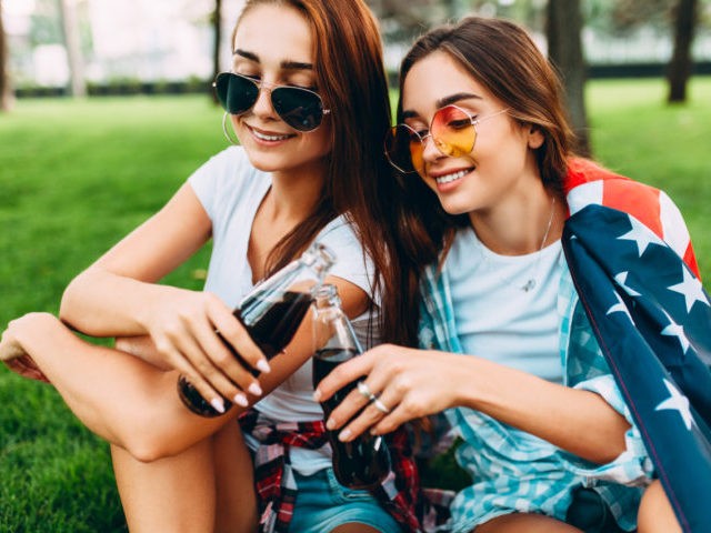 Two attractive young girls in sunglasses sitting in the Park on the lawn with the American flag, with a refreshing drink. U.S. independence day celebrations