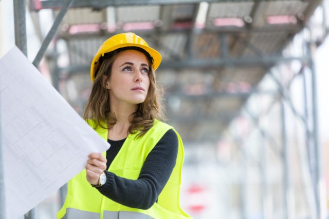Female construction worker with helmet and safety jacket on construction site examining office blueprints. Outdoors