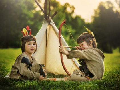 Cute portrait of native american boys with costumes, playing outdoor in the park with bow, arrows and hatchet on sunset, summertime