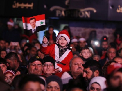 A young boy dressed in a Santa Claus outfit waves a flag with a portrait of Syrian Preside