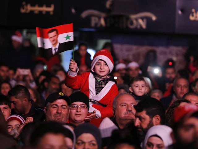 A young boy dressed in a Santa Claus outfit waves a flag with a portrait of Syrian President Bashar al-Assad as people gather in the centre of the Syrian capital Damascus to watch the lighting of a giant Christmas tree on December 23, 2018. (Photo by LOUAI BESHARA / AFP) …