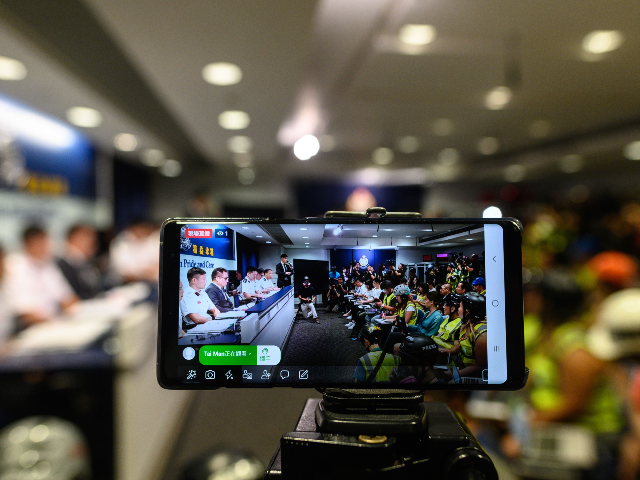 A live stream recording is seen on a mobile phone as journalists (R) wear protective gear