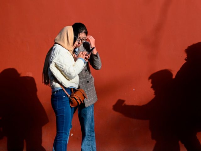Two women look at their mobile phone next to the shadow of two men taking a selfie at a pa