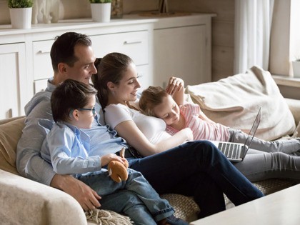 Happy parents hug kids lying on cozy sofa watching cartoons on laptop, young family spend time with children relaxing at home, mom and dad cuddle with son and daughter enjoying funny video at computer