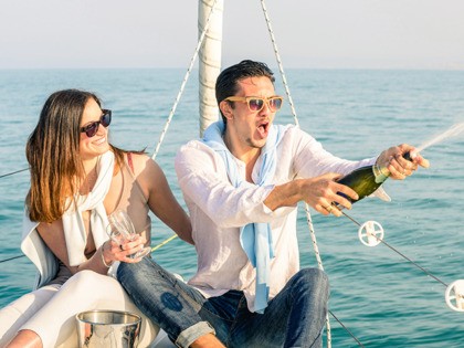 Young couple in love on sailing boat cheering with champagne wine bottle - Happy girlfriend birthday party cruise travel on luxury sailboat - Focus on boyfriend face with sunny afternoon color tones