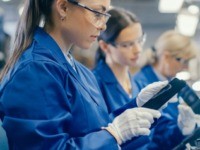 Woman Electronics Factory Worker in Blue Work Coat and Protective Glasses Takes a Smartphone Screen and Performs a Quality Check. High Tech Factory Facility with Multiple Employees.