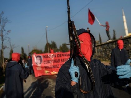A masked leftist militant holding an AK-47 walks in front of a banner reading "Revolution