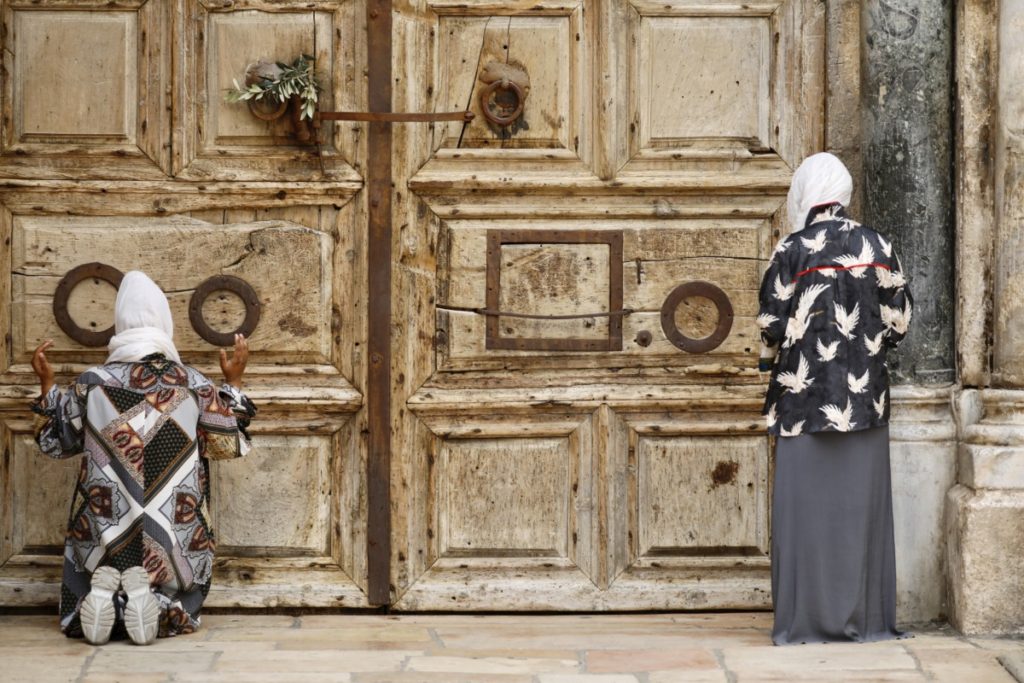 Women pray iin front of the closed Church of the Holy Sepulchre, a place where Christians believe Jesus Christ was buried, as a palm hangs on the door, in Jerusalem's Old City, Sunday, April 5, 2020. The traditional Palm Sunday procession was cancelled due to restrictions imposed to contain the spread of the coronavirus. (AP Photo/Ariel Schalit)