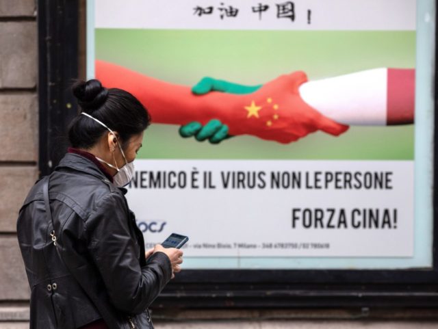 MILAN, ITALY - FEBRUARY 25: A woman, wearing a respiratory mask, walks past a placard showing a handshake between two hands representing the Chinese and the Italian national flags and reading 'The enemy is the virus, not the people. Go China!' in the Paolo Sarpi district (Milan's Chinatown) on February …