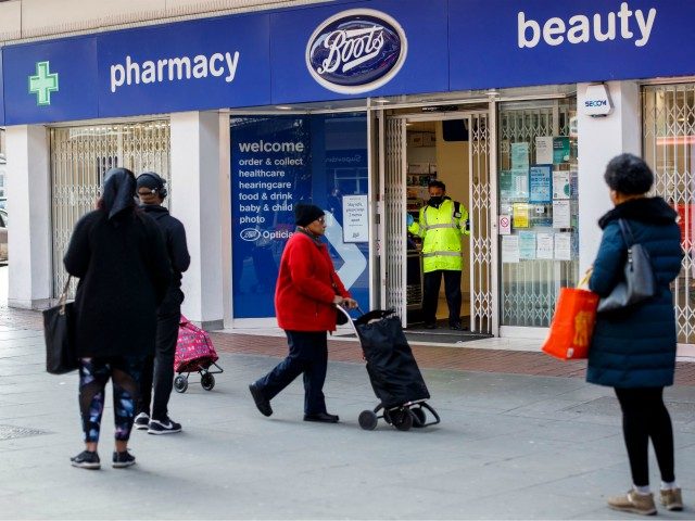 A security guard mans the doors of a Boots pharmacy as members of the public observe socia