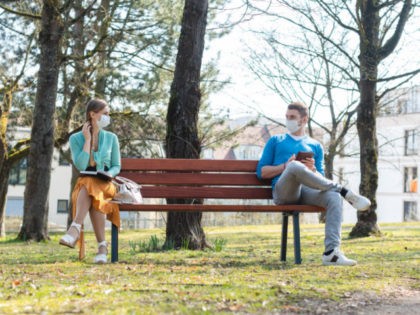 Woman and man with face mask in social distancing flirting sitting on a park bench