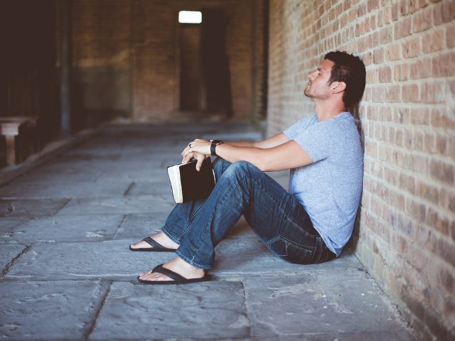 A man sits on the ground outside of a church with his Bible in his hands, apparently praying.