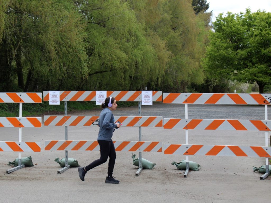 In this photo taken Wednesday, April 15, 2020, a woman jogs past a barricade outside a park in Salem, Ore. Oregon is in its fourth week of lockdown. Oregonians can't enter state parks in mountains and in valleys now blooming with springtime flowers, or go to the state's trademark wineries and microbreweries. But Oregon appears to be an outlier as coronavirus cases start to peak in each state. Of all the states in America, Oregon should have the fewest COVID-19 deaths per capita when the peak comes here, according to researchers at the University of Washington who developed a closely watched model. (AP Photo/Andrew Selsky)