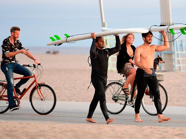 Surfers carry their boards as cyclists ride past, none wearing a facemask, at Santa Monica