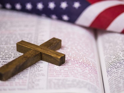 A holy Christian cross laying on an open Bible with an American flag in the background.