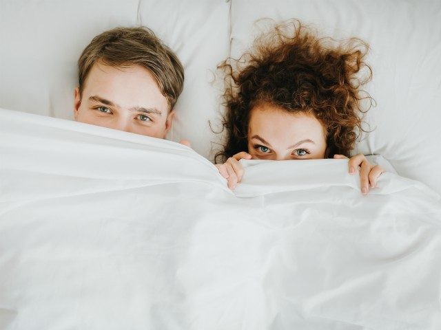 couple lying and hiding under white blanket - stock photo Happy fuuny couple lying and hiding under white blanket in bed