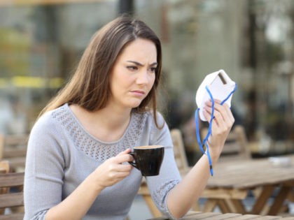 Doubtful young woman looking suspicious at protective face mask on a coffee shop terrace