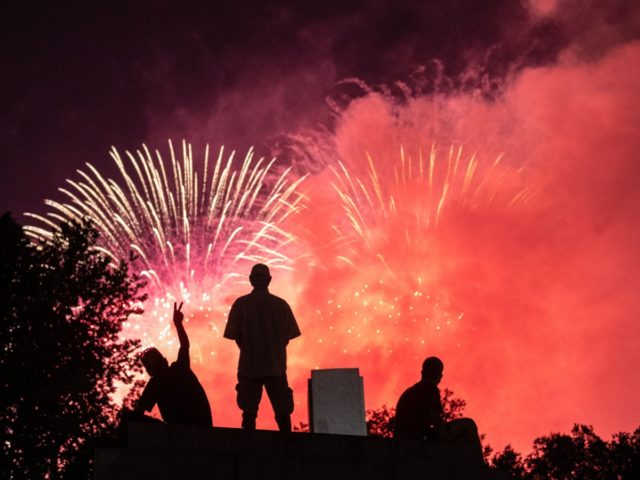 Spectators sit on top of a pillar at the World War II Memorial in the Mall as fireworks bu