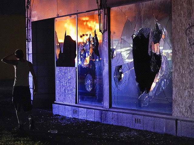 A protester walks past a building with broken windows as others burn during protests, Mond