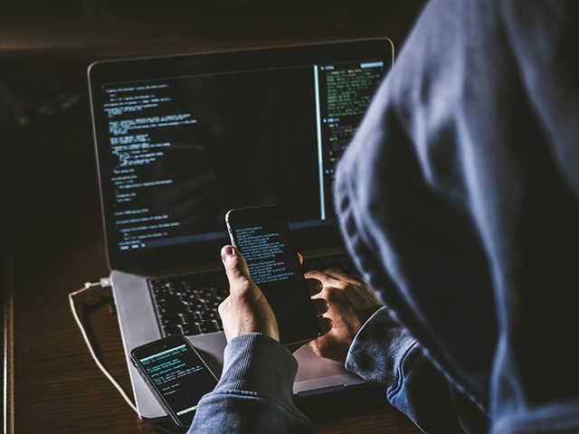 anonymous person in the hood sitting in front of computer working with laptop and mobile p