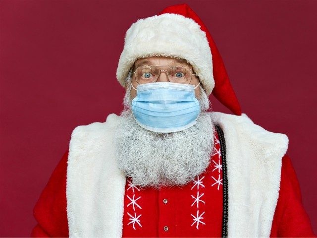 Close up portrait of funny old bearded surprised Santa Claus wearing costume, glasses, face mask looking at camera standing on Christmas red background. Covid 19 coronavirus safety protection concept. - stock photo