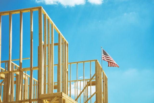 American flag waving in the wind on the top of a construction home framing. Blue sky and w