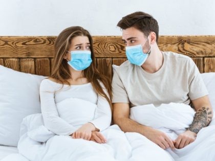 Disease and quarantine concept. Sad guy and girl in protective masks are sitting in bed and looking at each other